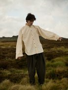 Person wearing a beige shirt and dark pants standing in a grassy field with a cloudy sky.