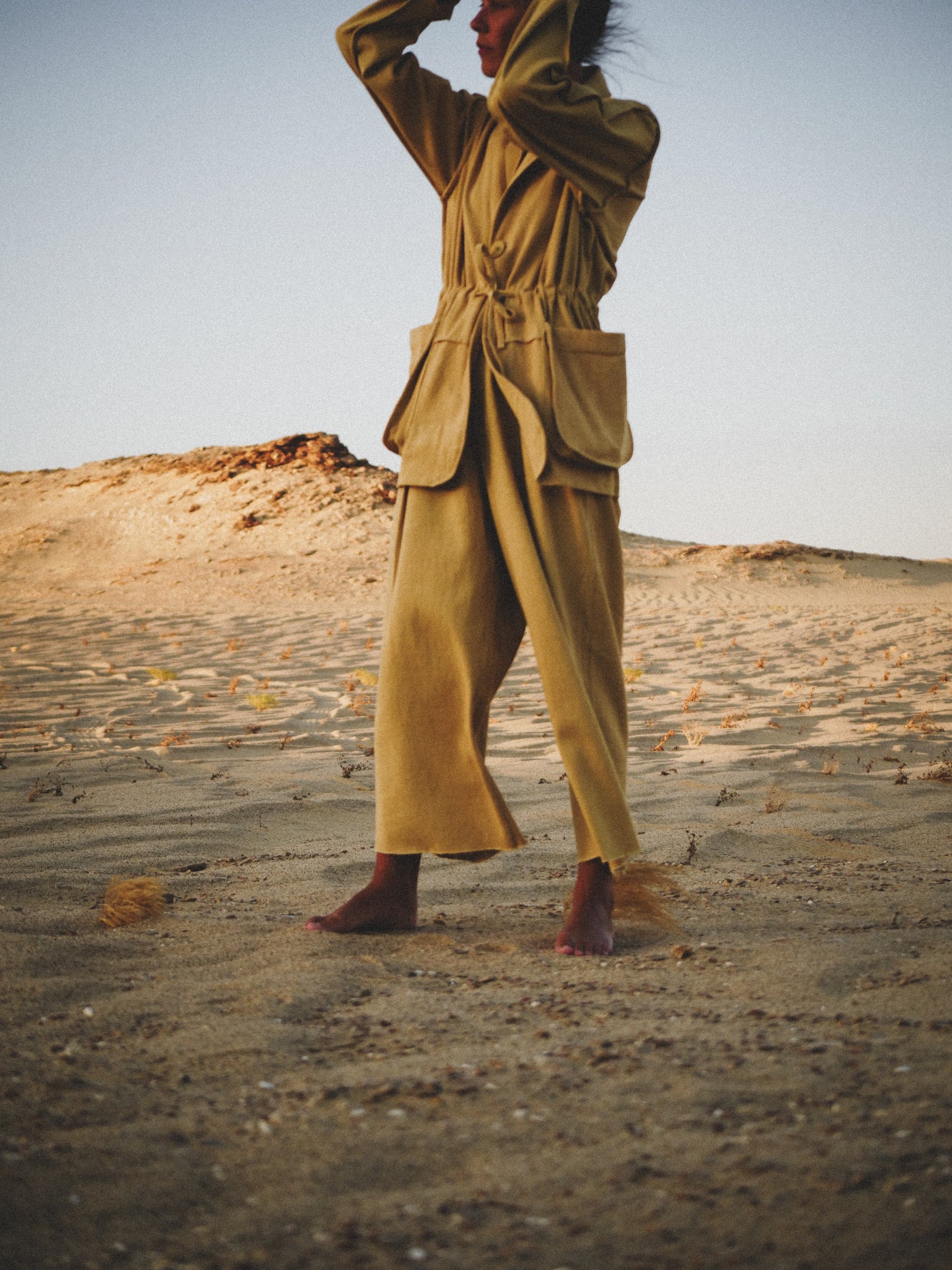 Person wearing a pale mustard jacket and pants standing in a desert landscape.