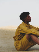 Person wearing a mustard yellow outfit sitting on sand with a neutral background