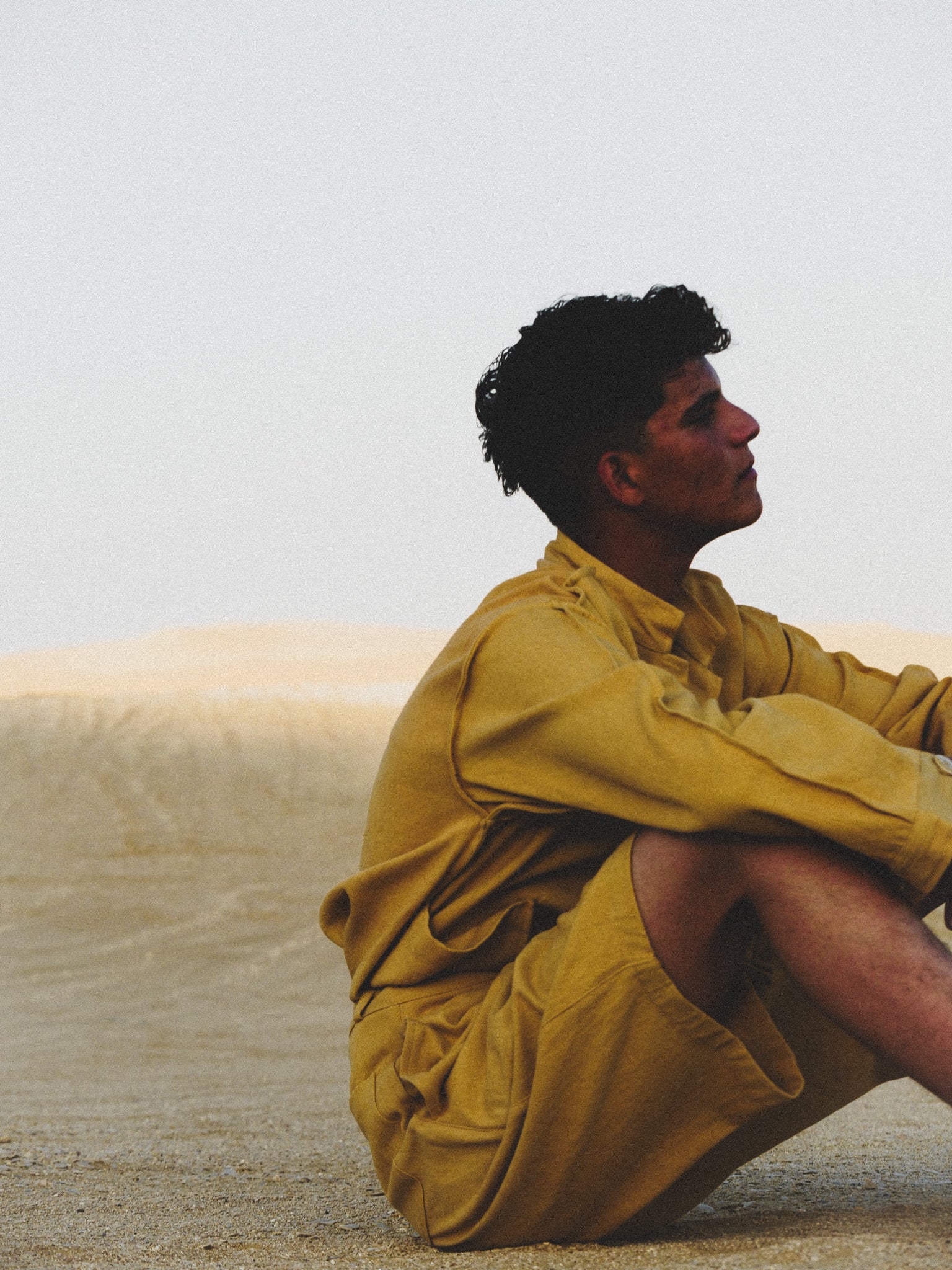 Person wearing a mustard yellow outfit sitting on sand with a neutral background