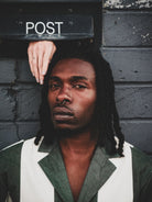 Person with braided hair wearing a green and white jacket against a textured wall with a 'POST' box.