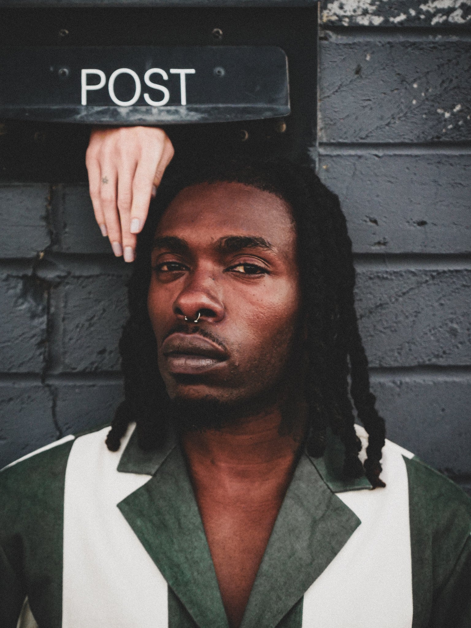 Person with braided hair wearing a green and white jacket against a textured wall with a 'POST' box.
