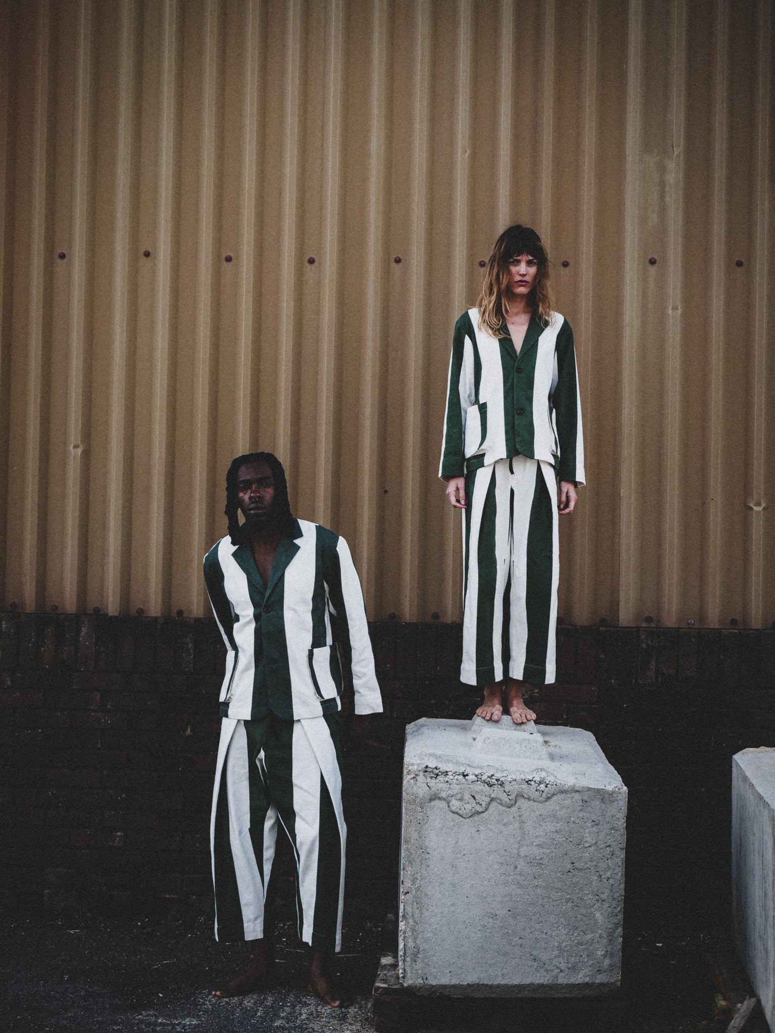 Man and woman wearing green and white outfits against a corrugated metal wall.