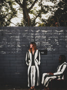 Two people in black and white outfits against a dark brick wall with trees in the background.