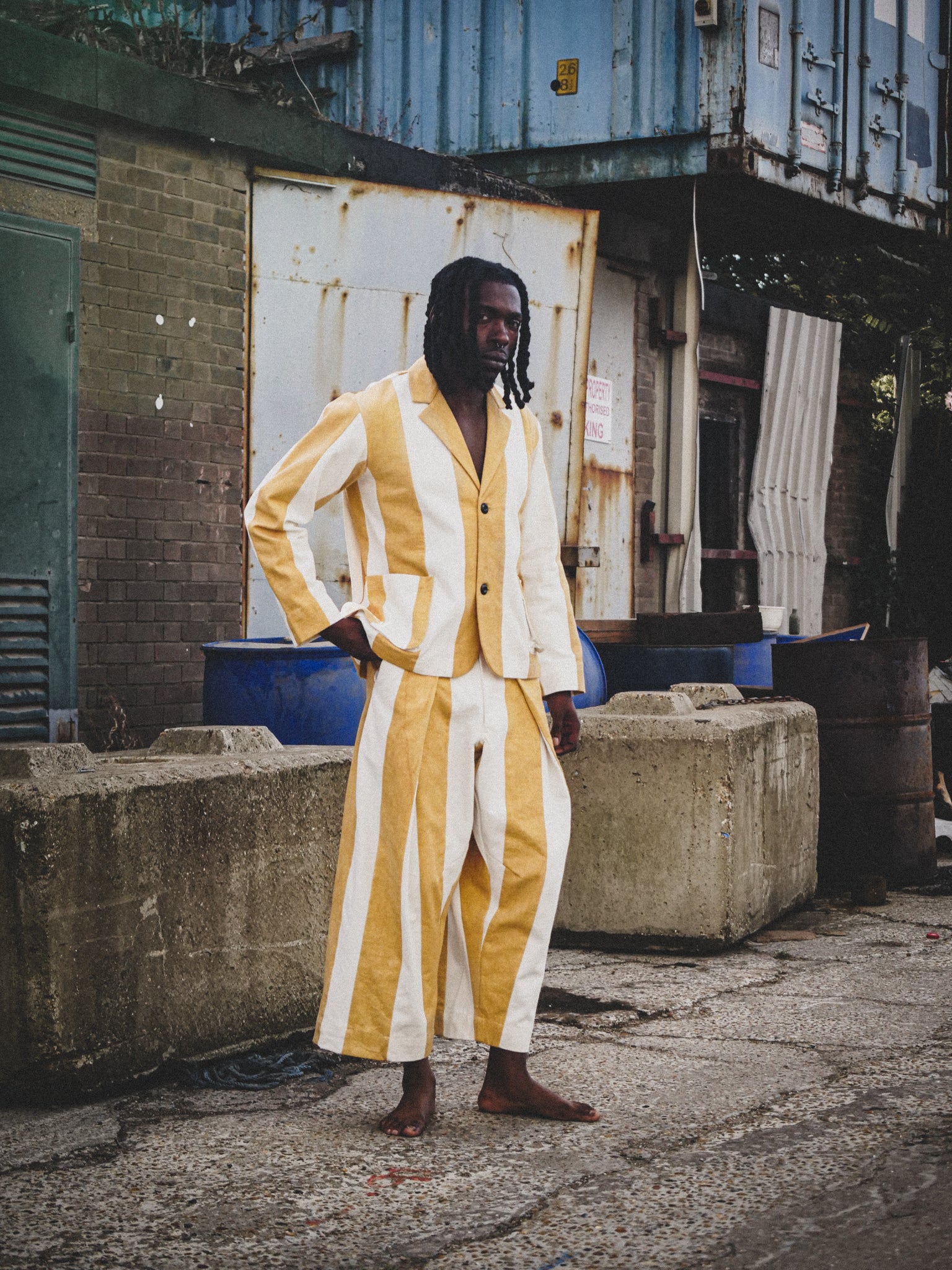 Man wearing a mustard and white striped suit standing in an urban setting with industrial background.