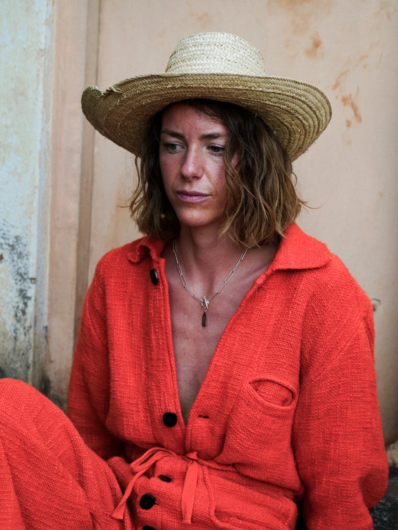Woman wearing a red outfit and straw hat against a textured wall.