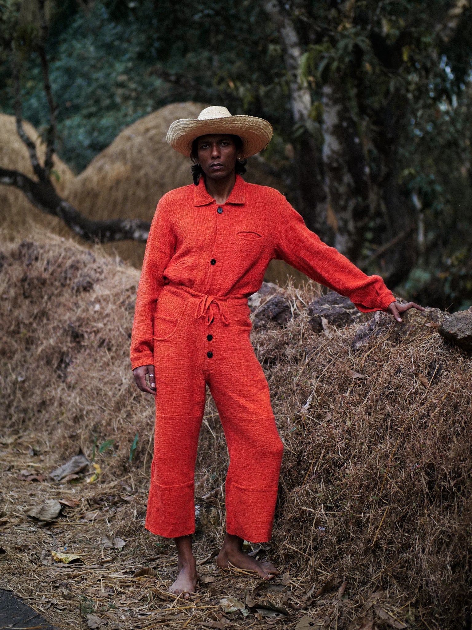 Man in sombrero wearing an orange jumpsuit and straw hat standing on a pile of hay with a natural background
