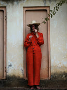 Woman in sombrero wearing an orange jacket and trousers and straw hat standing in a doorway.