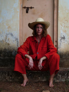 Woman in sombrero wearing orange jacket and trousers sitting against a weathered wall.