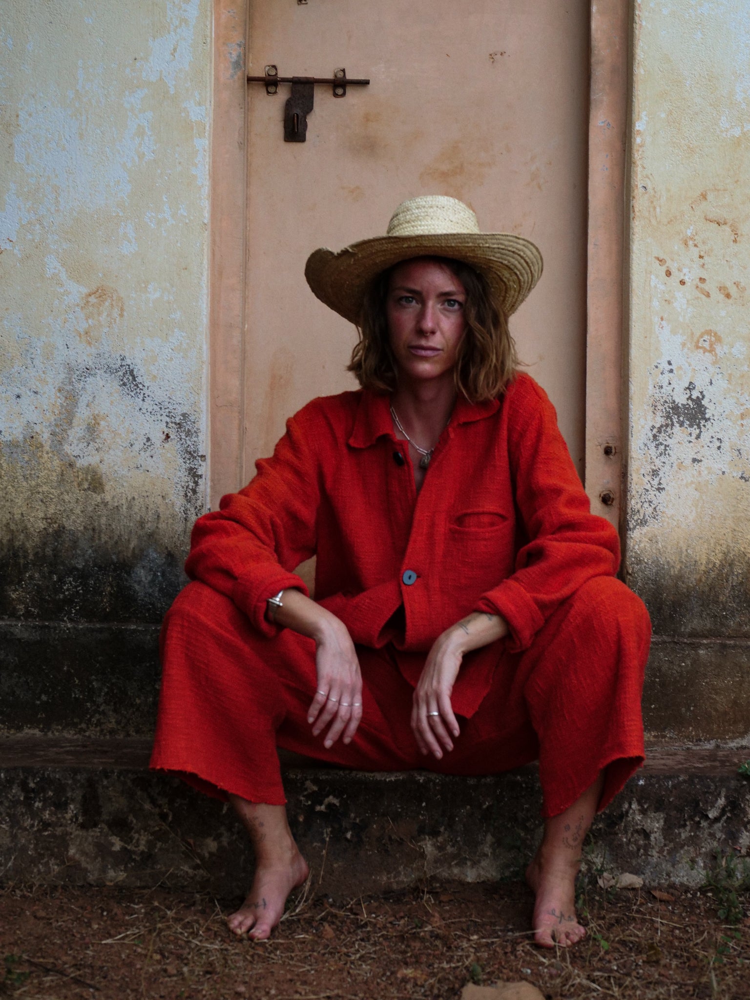 Woman in sombrero wearing orange jacket and trousers sitting against a weathered wall.