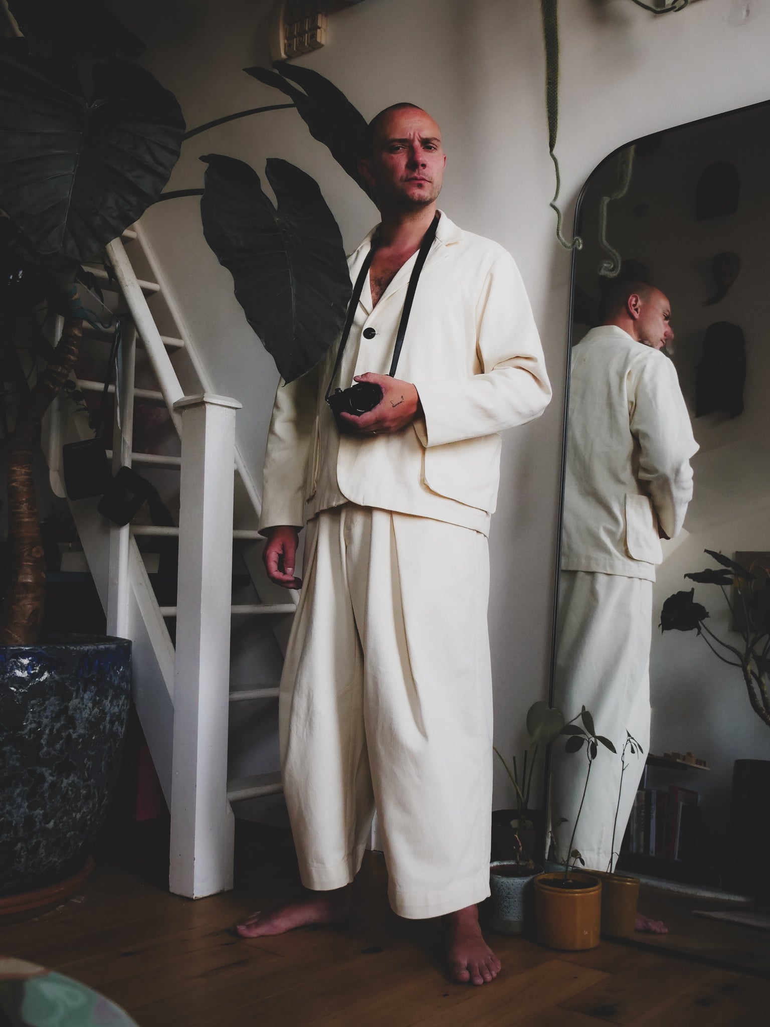 Man in a white suit standing in a room with plants and a staircase.