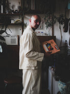 Man in a cream-colored outfit holding a colorful box in a room with plants and shelves.