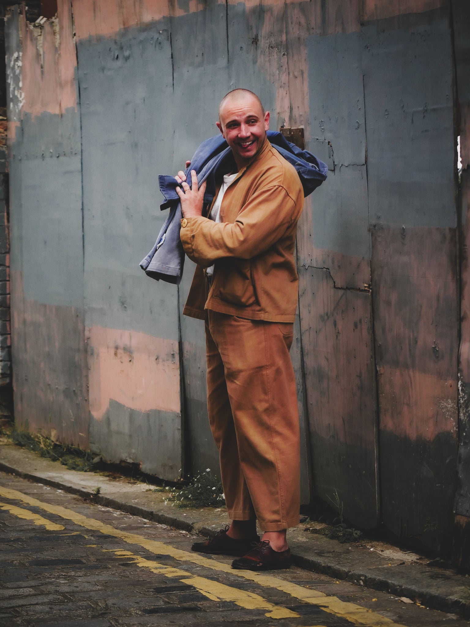 Man in a brown suit holding a blue jacket against a textured wall.