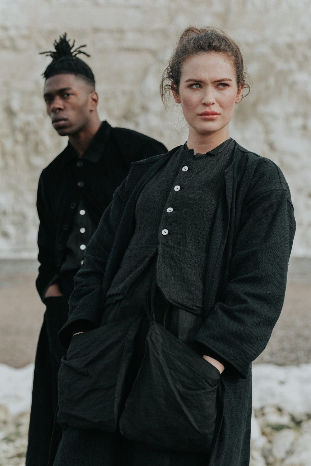 Two people wearing black outfits standing against a textured wall.