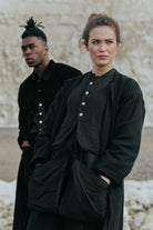 Two people wearing black outfits standing against a textured wall.